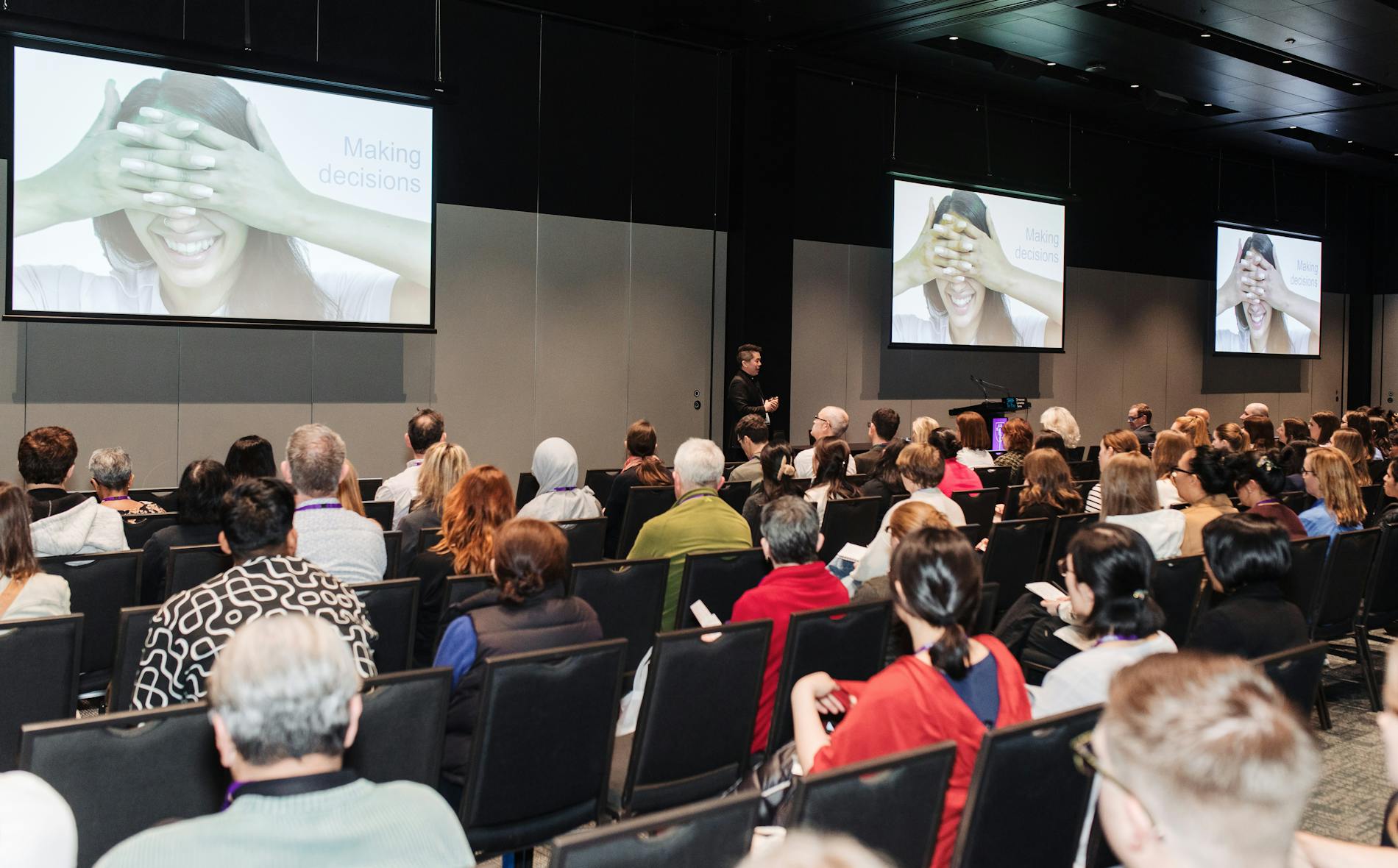 A speaker presents at a conference, discussing "Making decisions," while an audience attentively watches. Two large screens display the Te Pae branding and relevant visuals emphasizing the topic.